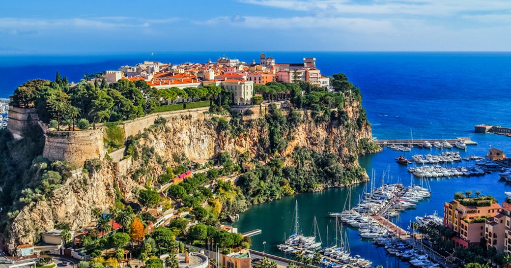 Elevated view looking over Monaco below, multiple yachts moored in marina with settlement upon cliff top and sea in the distance.