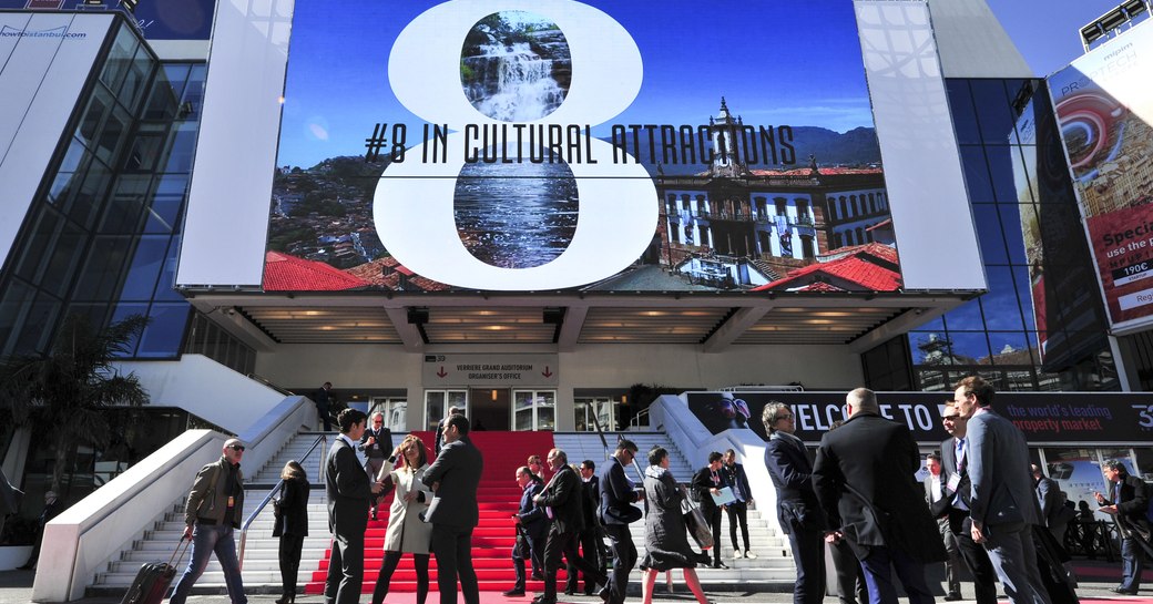 Red carpet entrance to MIPIM exhibit, with big screen overhead and visitors gathering outside.