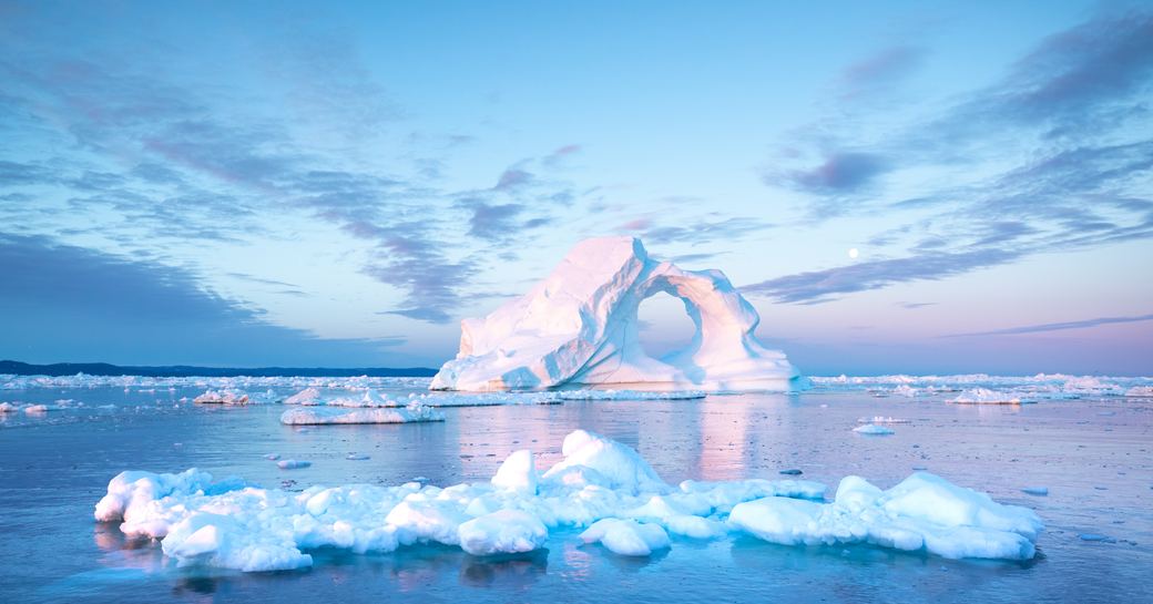 Intricate iceberg with a hole under an interesting and colorful sky during sunrise with full moon.