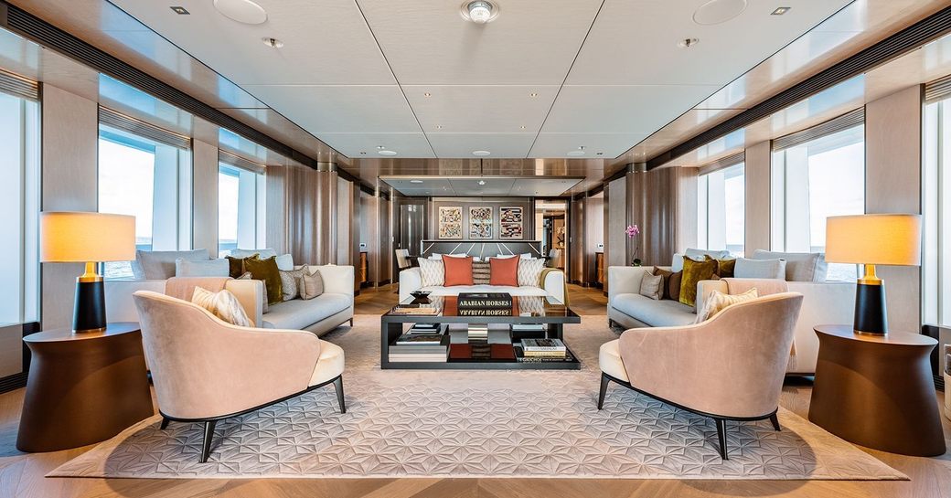 Overview of the main salon lounge area with cream armchairs and sofas surrounded by large windows onboard charter yacht MOSKITO