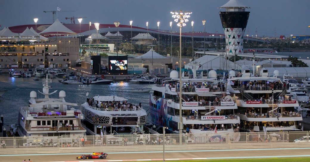 Car races along track at Abu Dhabi Grand Prix circuit at night, with superyachts lined up along the dock