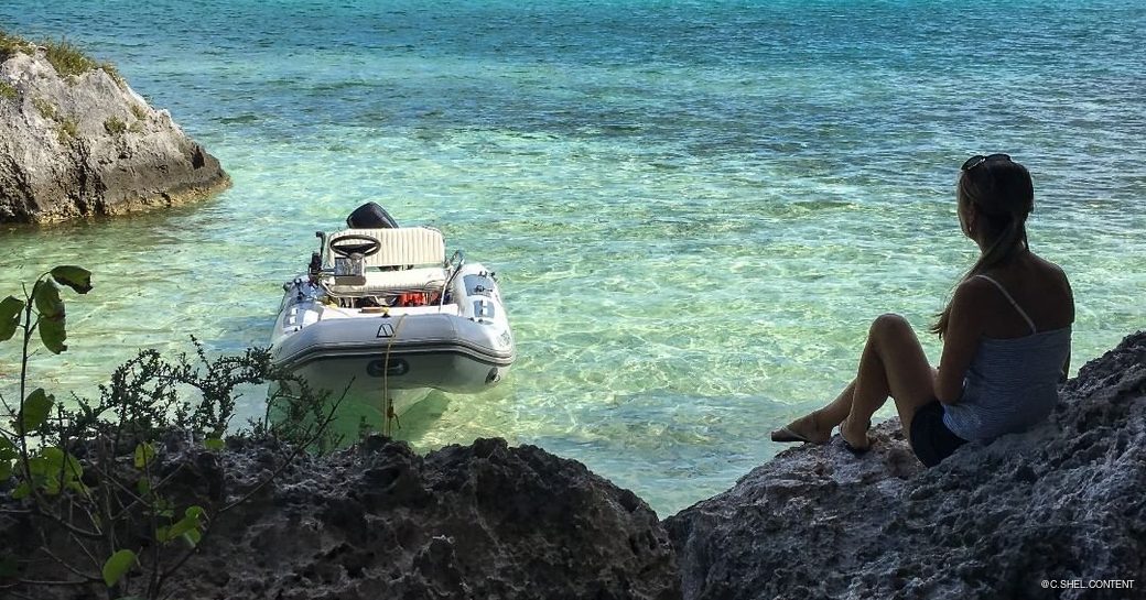 Girl sat on rocks in the shade looking out at boats anchored in blue waters 