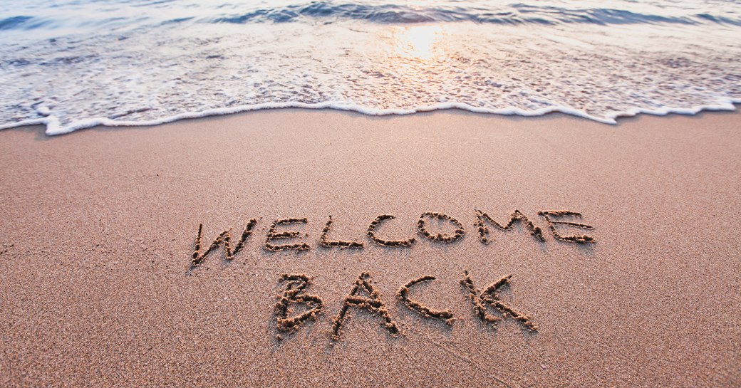 "Welcome back" written in caps in the sand on a beach lapped by waters at sunset