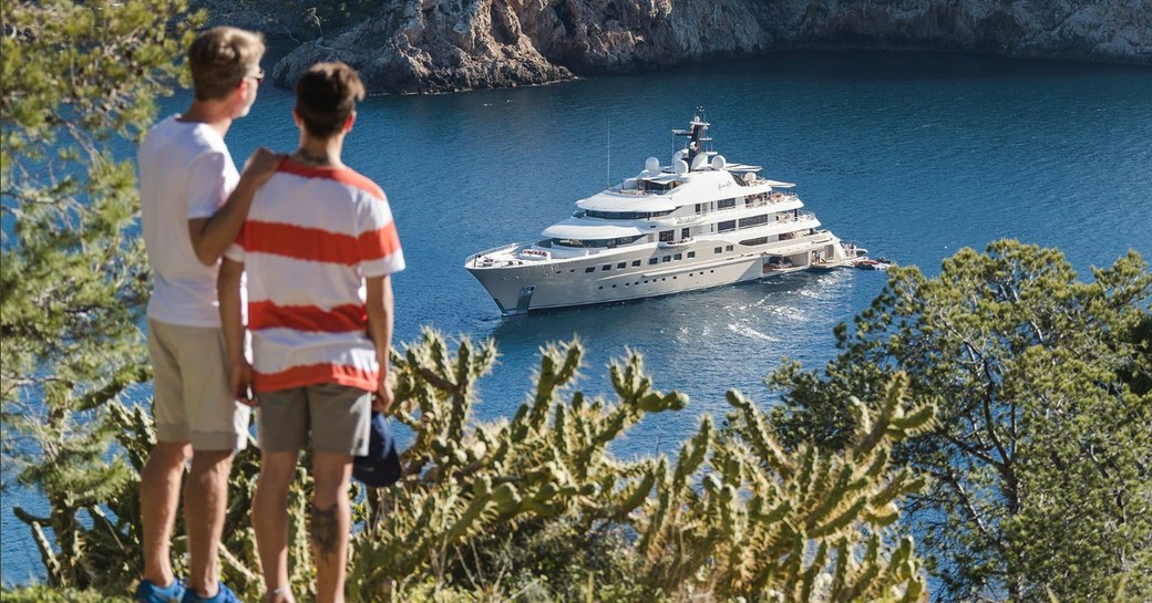 two young men stand on cliff looking over superyacht Here Comes The Sun anchored in water 