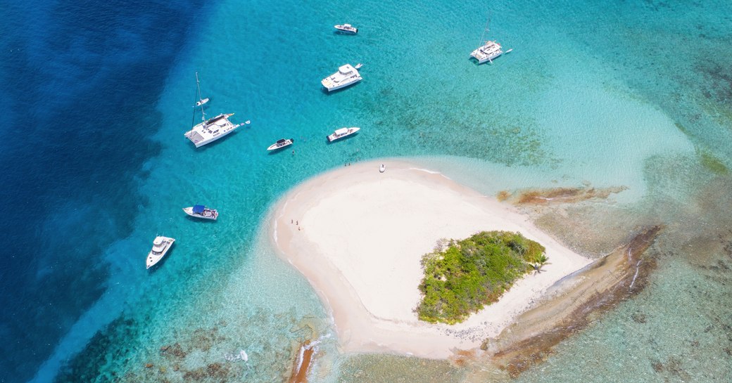 Sand island with small crop of trees viewed from above and yachts in clear water around it