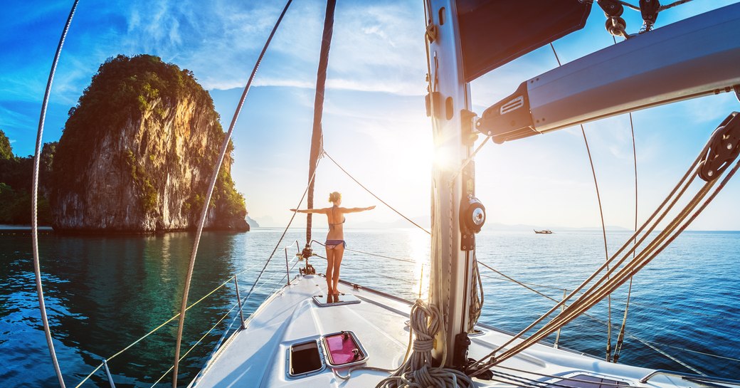 A woman stands with arms outstretched on the bow of a sailing yacht in Thailand