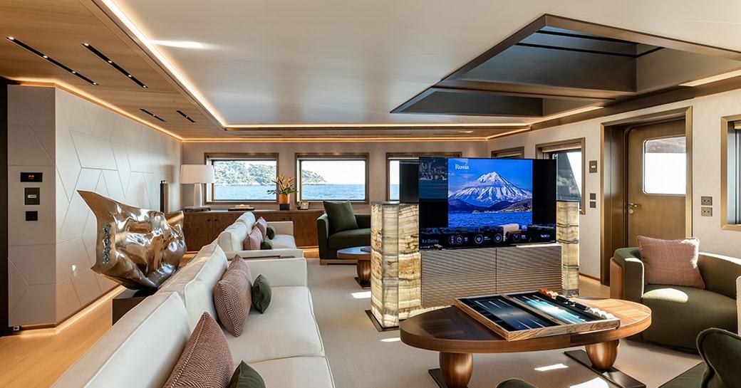 Overview of the main salon with plush sofas and a large TV screen surrounded by windows onboard charter yacht LA DATCHA