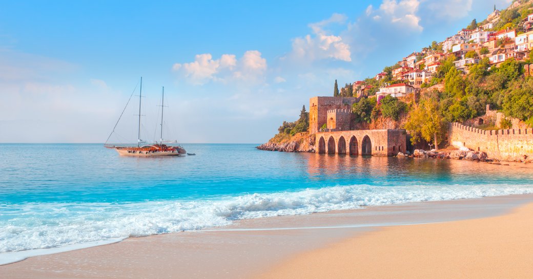 A gulet anchored in blue waters opposite a steep hill nestled with houses leading down to a medieval fortress