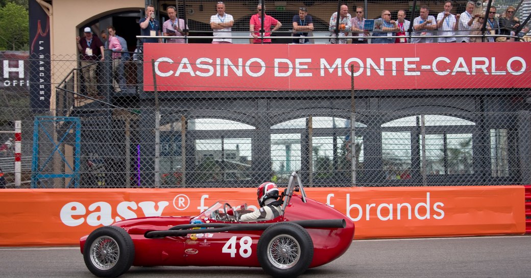 Racer in vintage red car at Monaco Historic Grand Prix, with Casino de Monte-Carlo sign in background with spectators watching from terrace.