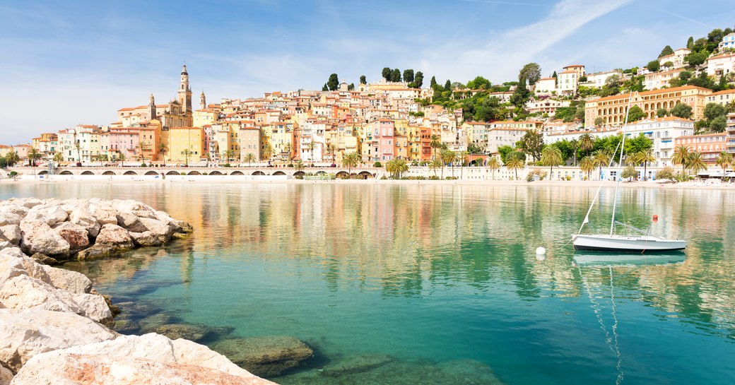 yacht anchored in clear waters with rocks and colourful french village in the background