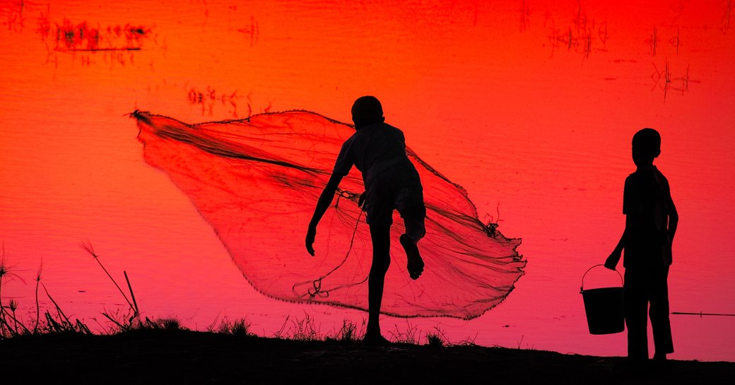 Two young fisherman seen from behind as they cast out a net on water reflecting the sunset