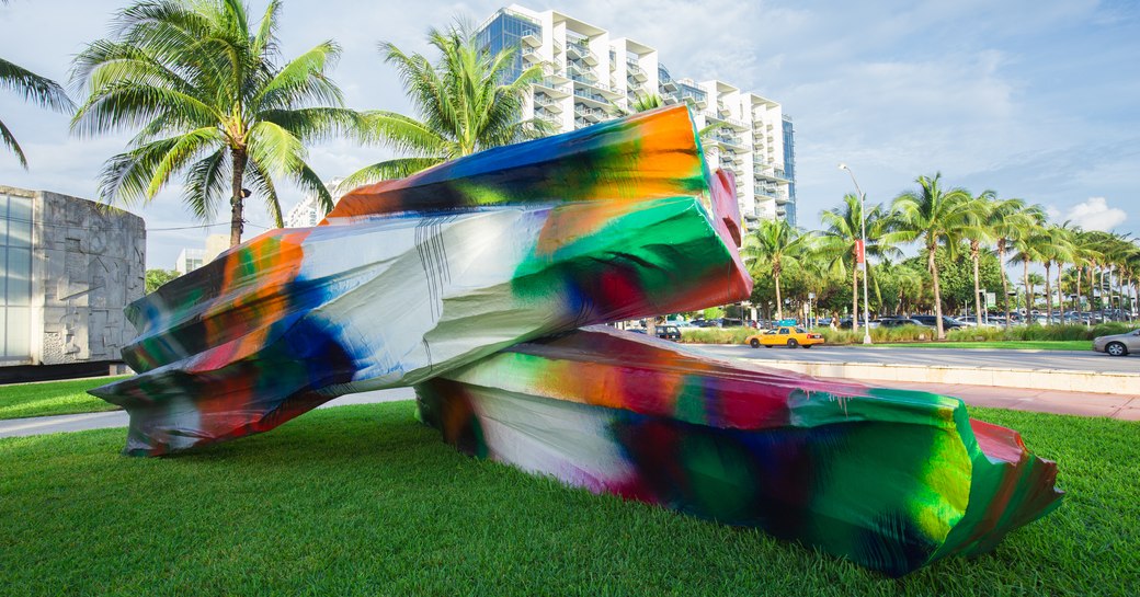 Sculpture on display during Art Basel Miami, rainbow colours shine out against green grass and towering Miami hotels in background.