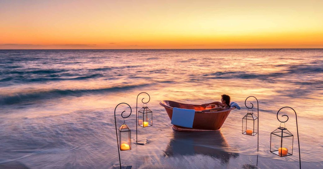 woman relaxes in copper bath on the beach surrounded by lanterns on thanda island