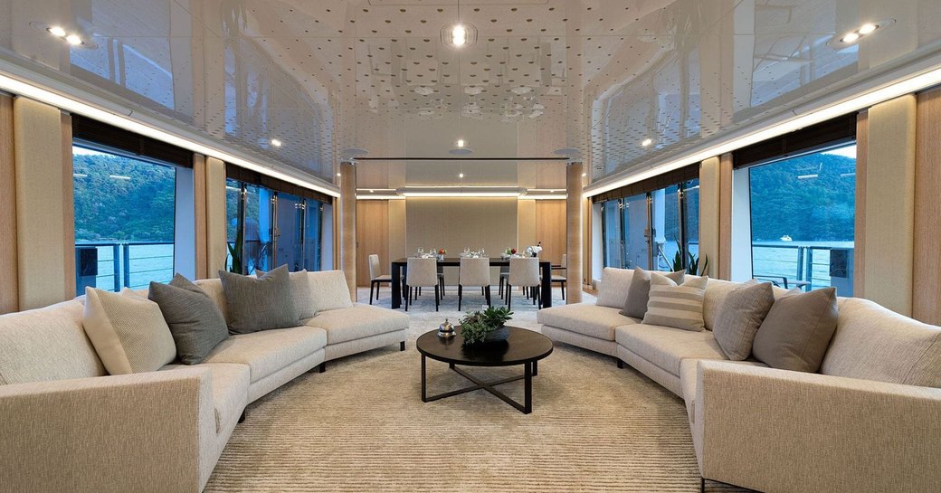 Overview of the main salon onboard charter yacht AQUARIUS, large lounge in the foreground with surrounding full height windows, with dining area aft.