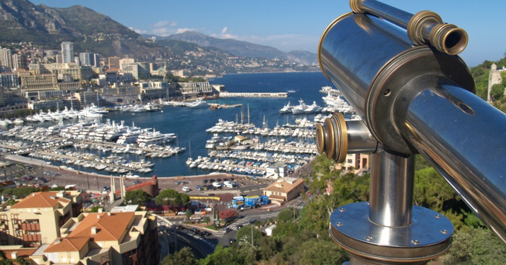 Elevated view of Monaco marina, telescope in foreground overlooks Monaco from hill top, with buildings cascading down to the marina. 