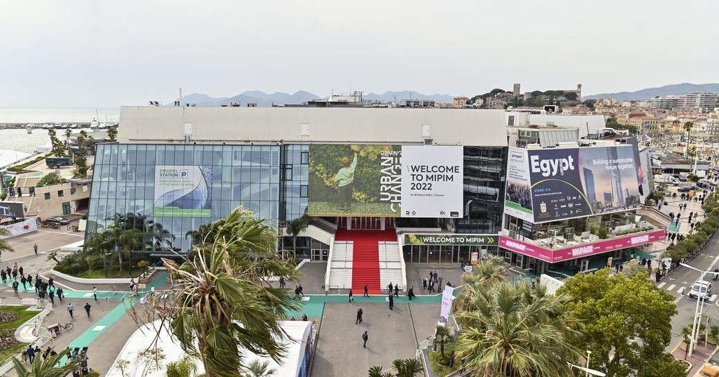 Exterior view of Palais des Festivals, Cannes, during MIPIM real estate event. Red carpet visible with small gathering of visitors exploring.