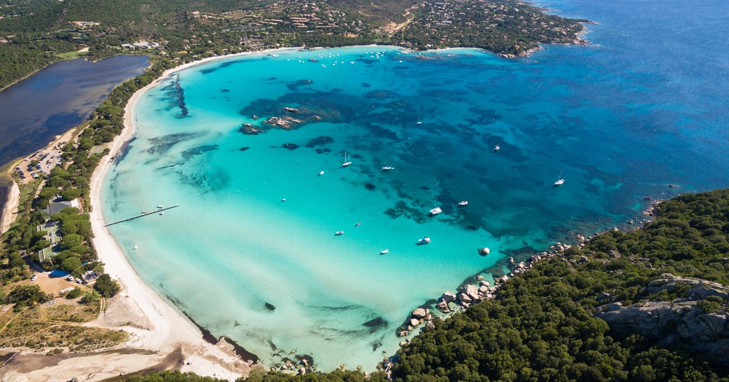 Aerial view of bright blue water in Corsica, with boats dotting the water and strip of sandy beach backed by green landscapes