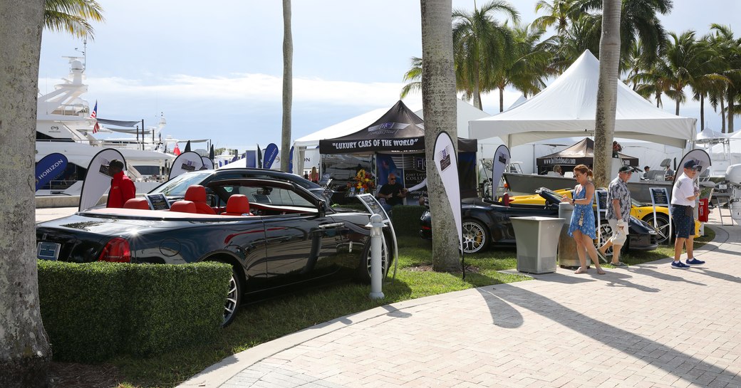 Super cars on display along a walkway at the Palm Beach International Boat Show. Superyachts visible in background, with visitors looking at the cars.