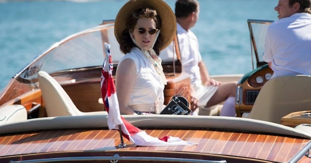 Actor in the Crown playing Queen Elizabeth looking back with sunglasses and a hat on onboard a luxury tender
