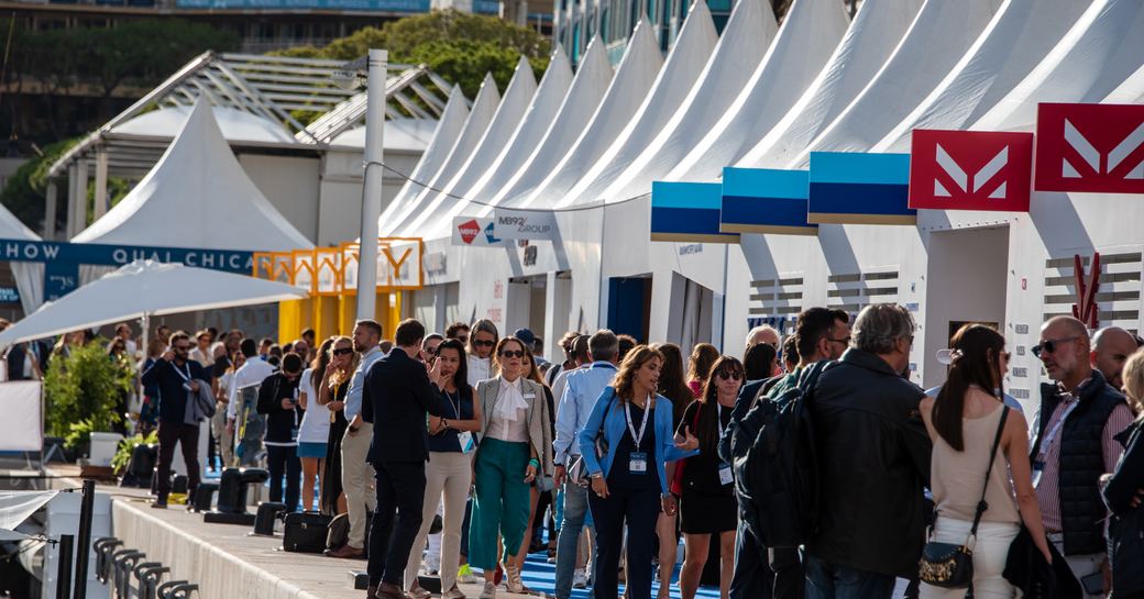 Line of exhibitor stands at the Monaco Yacht Show with visitors walking by.