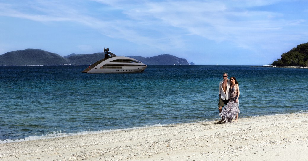 A couple walk hand in hand on the beach whilst superyacht 'Ocean Emerald' sits at-anchor a short distance away