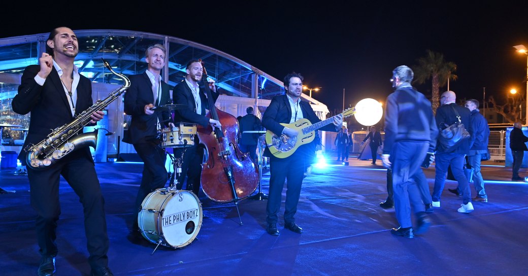 Frontal view of live band at MIPIM event, musicians playing drums, guitars and saxophone.