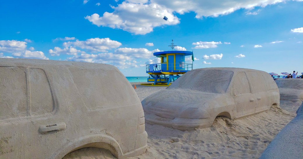 Landscape image of Art Basel Miami sand sculptures, cars etched into sand representing traffic.