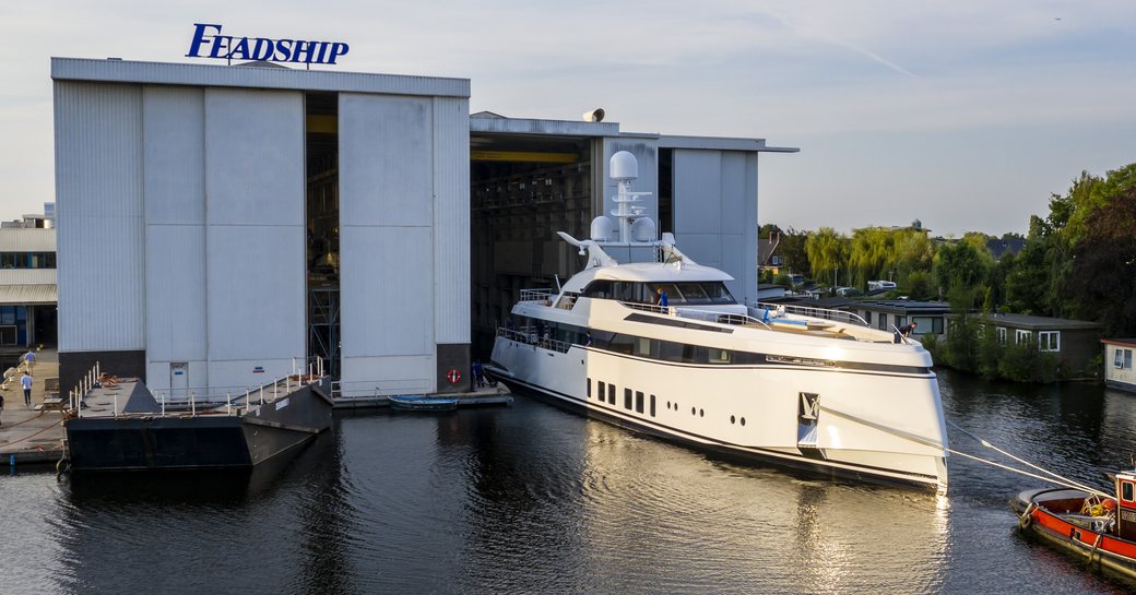Feadship superyacht Project 706 leaving Facilities at Aalsmeer, with Feadship branded building behind on calm water