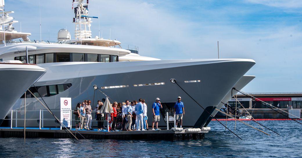 Group of visitors attending the Monaco Yacht Show standing on a pontoon next to the bow of a superyacht