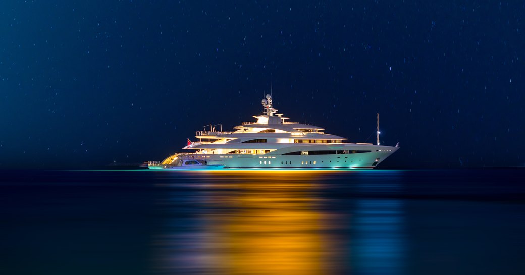 Night view to large illuminated white boat located over horizon, colorful lights coming from yacht reflect on the surface of the the Gulf sea.