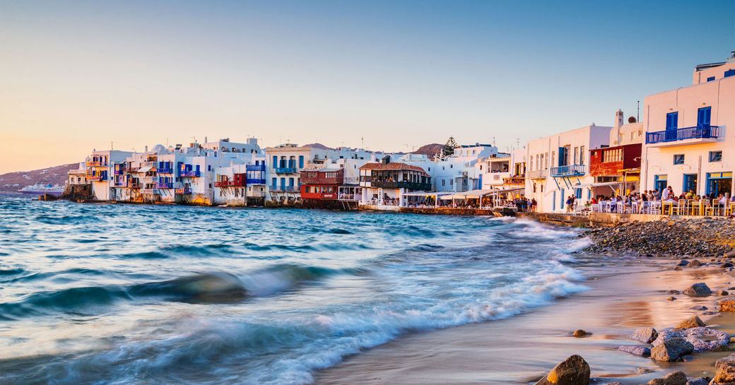 The sea hitting the shore of Mykonos, pastel buildings can be seen in the background