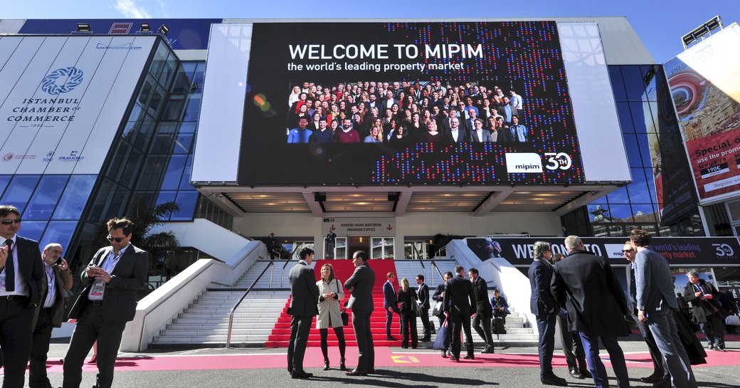 MIPIM posterboard displayed at Palais des Festivals, Cannes. Visitors grouped outside in discussion.