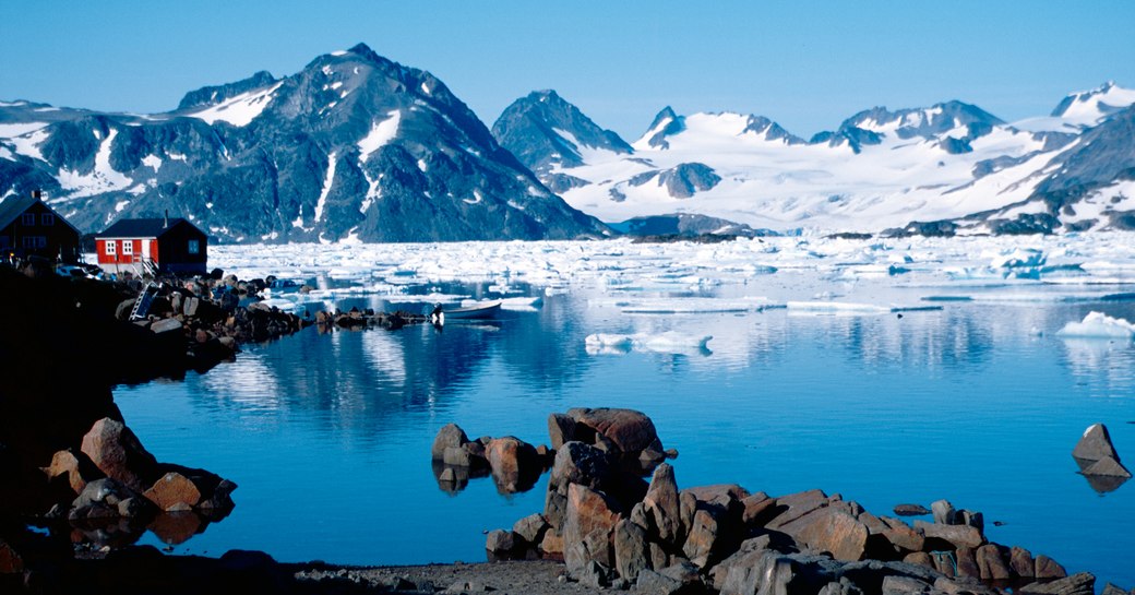 Overview of a seascape in Greenland, with a small red house on the left and snowy mountains in the background