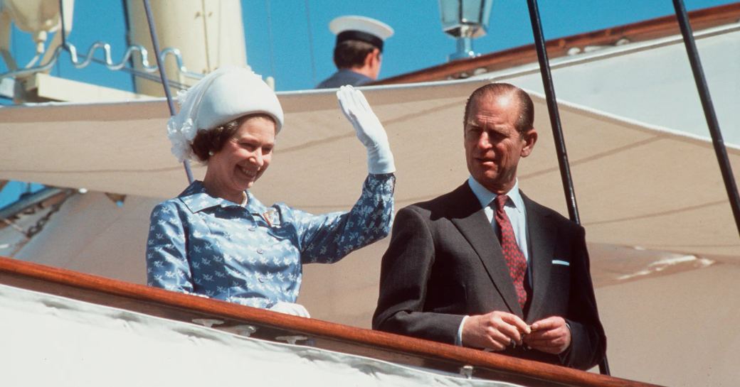 Queen Elizabeth waving to crowds onboard Royal Yacht Britannia with Prince Phillip by her side