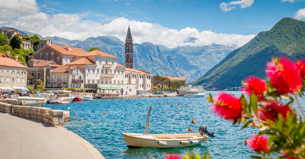 Seascape of marina in Montenegro, with flowers in foreground and mountains in background with town with clay red rooves in centre of shot