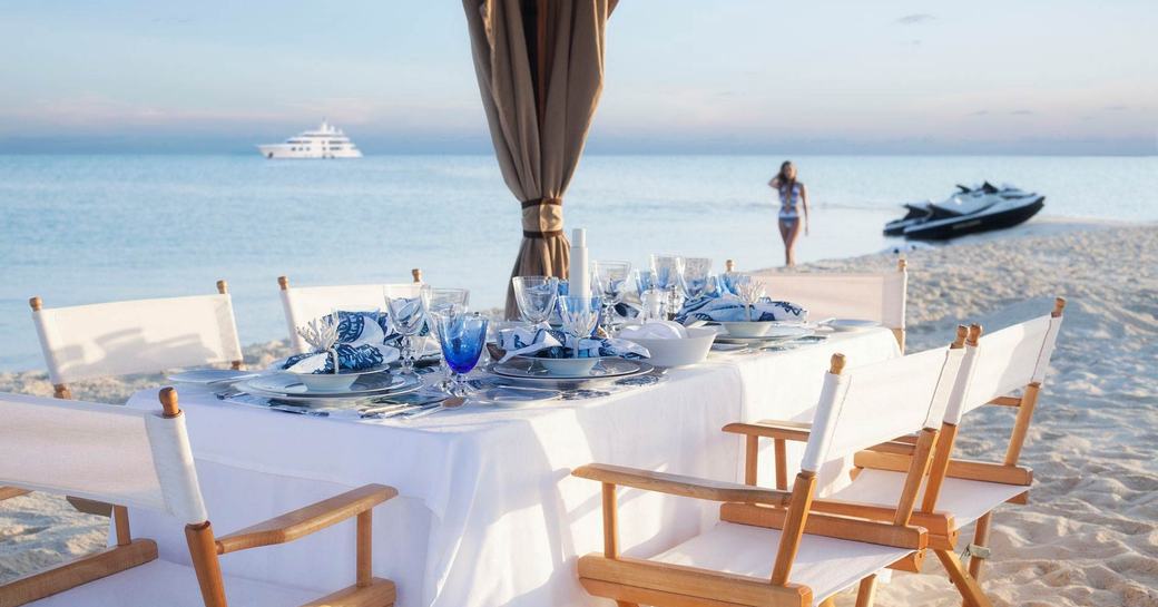 Dining table set up on beach with female walking towards it near tender boat and large superyacht on sea  in the distance