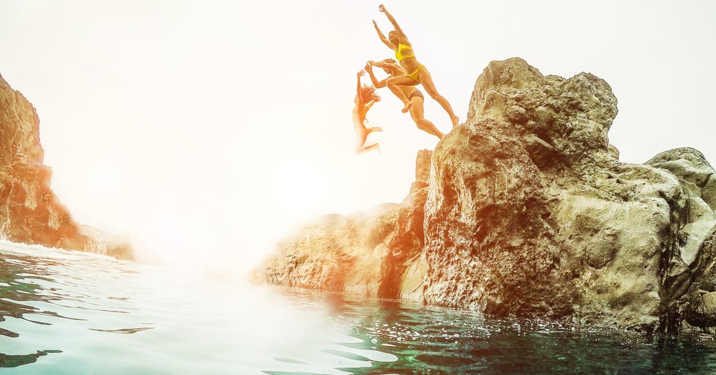 Three youths jump off a rock set in aquamarine waters against a bright sun in Ibiza in the Balearics, Spain