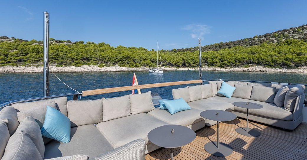 Exterior lounge area onboard charter yacht JICJ, grey seating with three coffee tables and view of a lush, green bay in the background