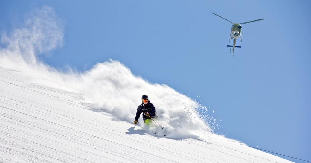 A charter guest skiing down a snowy slope with a helicopter in the sky above