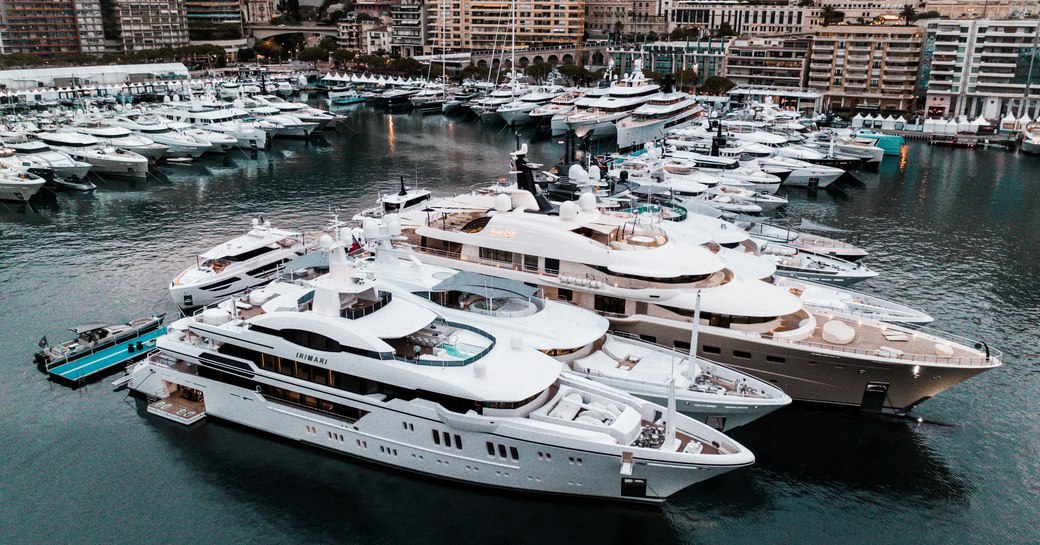 Superyachts moored along jetty at Monaco Yacht Show, Monte Carlo in background.