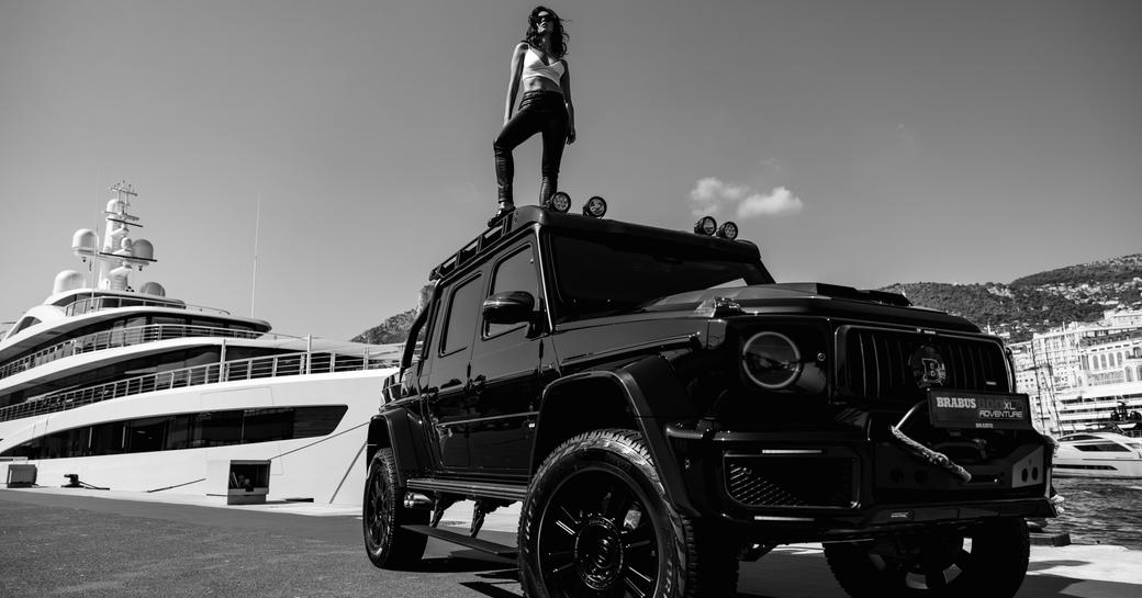 A black and white image of a woman standing on an all-terrain vehicle with a superyacht in the background 
