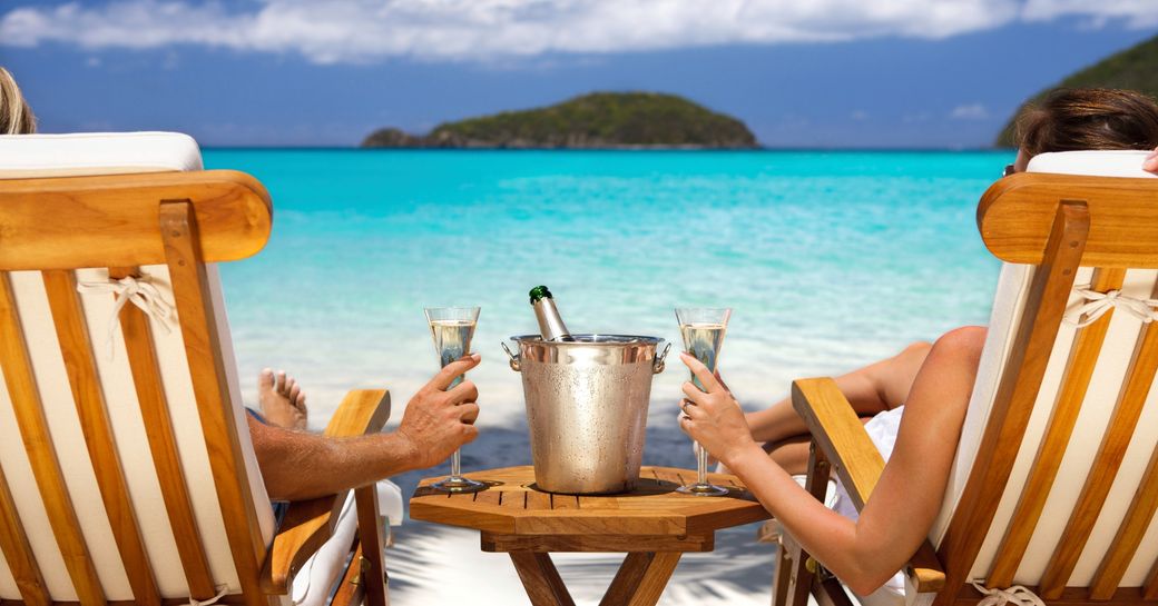 An unseen couple sit on deckchairs facing the blue Caribbean waters and islands with a bucket of champagne and two glasses between them