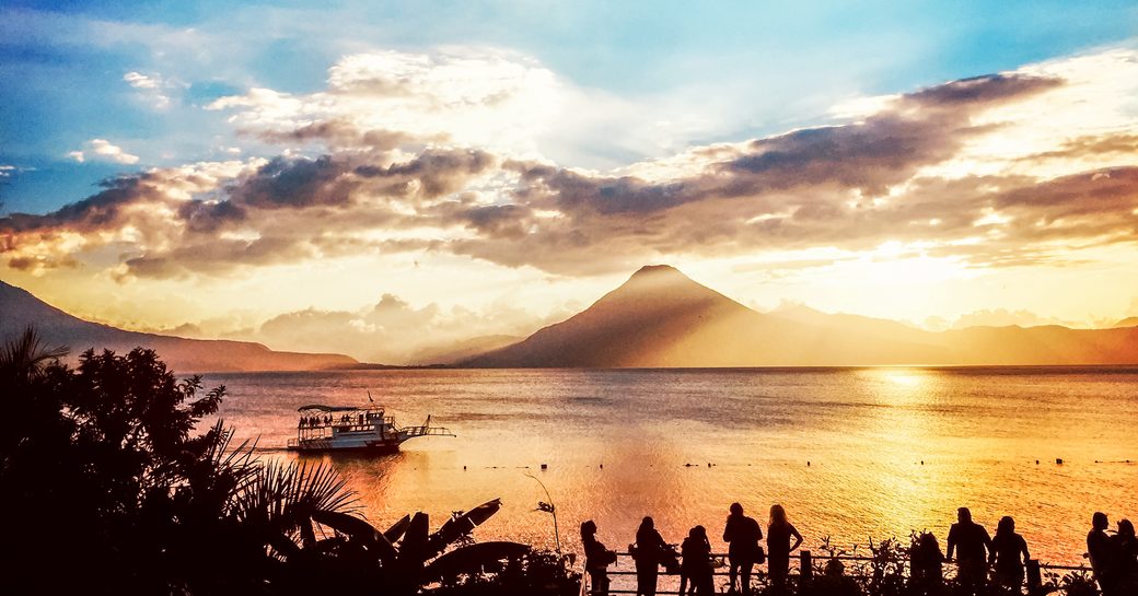 Silhouetted tourists watch sunset over Lake Atitlan & San Pedro volcano as ferry boat cruises past, Guatemala, Central America