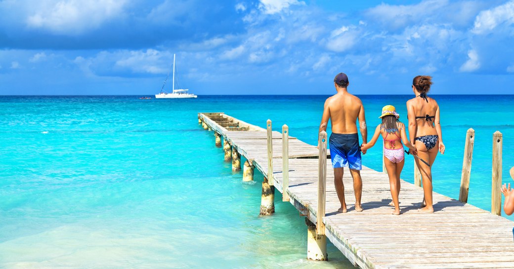 A family stroll down a wooden pontoon over a turquoise sea with a sailing yacht in the distance