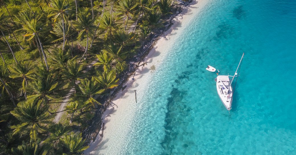 white Sailing Yacht anchored in crystal clear Turquoise Water right next to the perfect White Sand Beach of a Tropical Island in the Caribbean