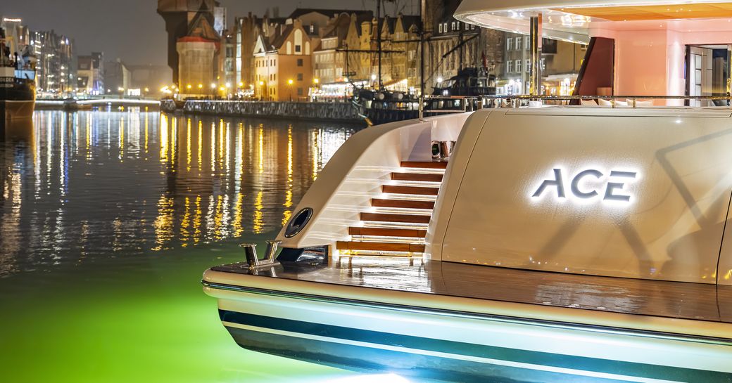 Aft view of the swim platform and nameplate of charter yacht ACE, surrounded by water lit by underwater lights and a marina lit up in the dark.