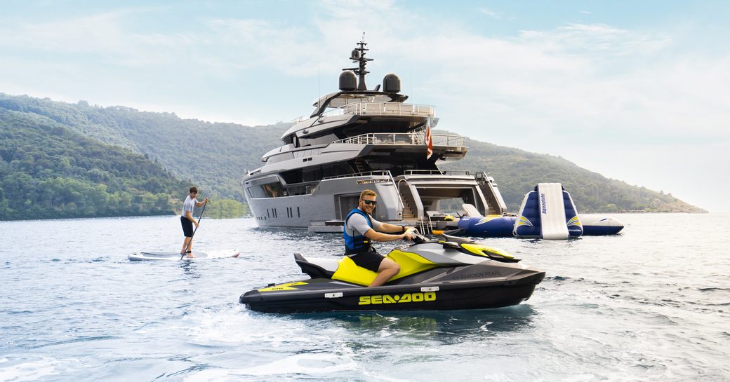 Aft view of charter yacht PANDION PEARL anchored at sea, with charter guests using water toys. Paddleboarder to port of superyacht with man on Jet Ski in the foreground. 