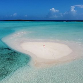 Drone shot of woman standing on sand surrounded by blue water 