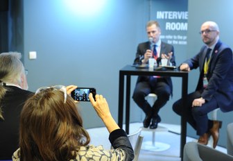 Two men on stage with microphones at MIPIM, with woman in the audience taking a photo