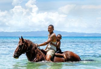 A man and his child on horseback wading through the shallow sea water in Fiji